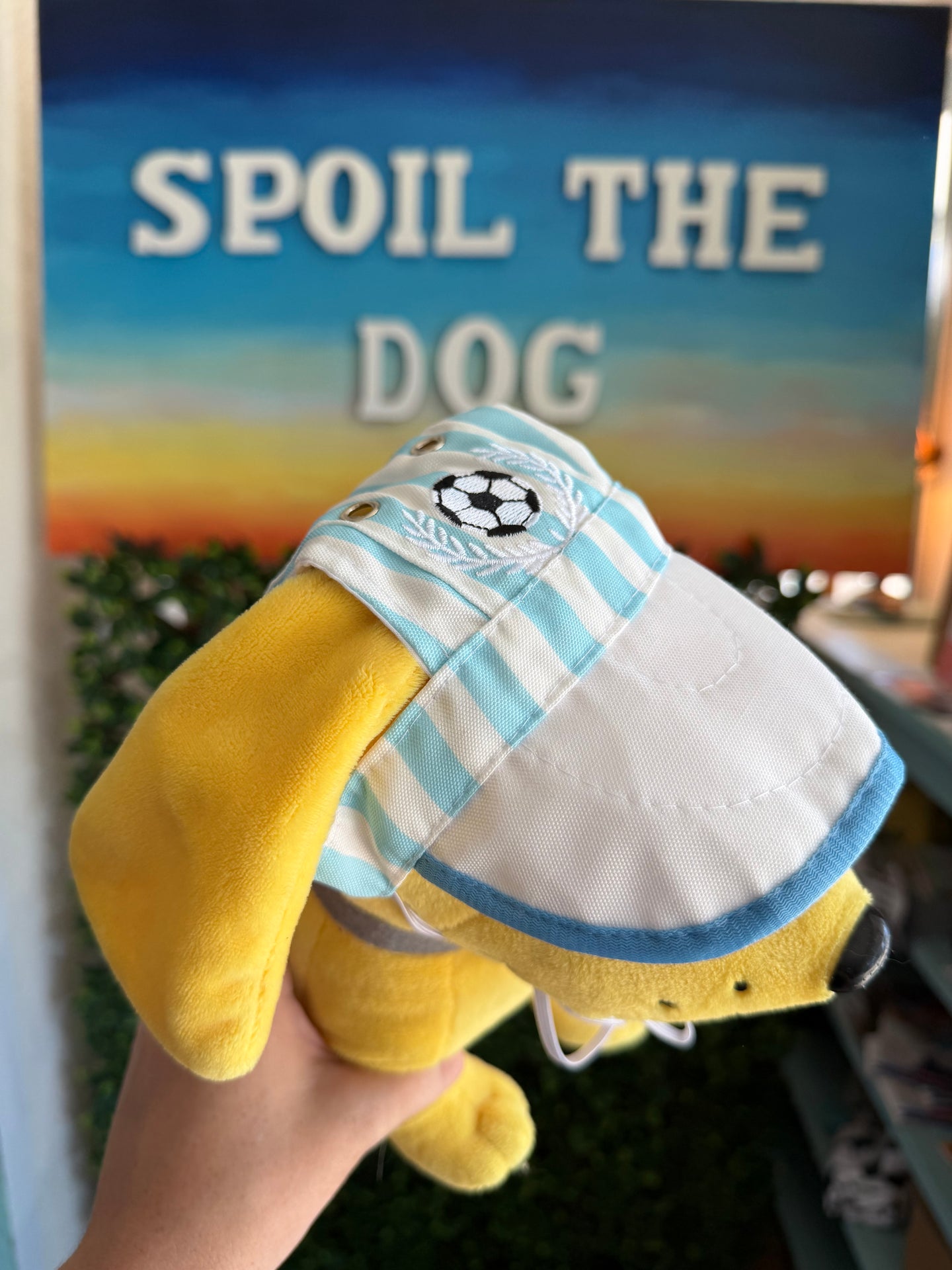 Yellow plush dog toy with a striped shirt held in front of a sign that says 'Spoil the Dog'.