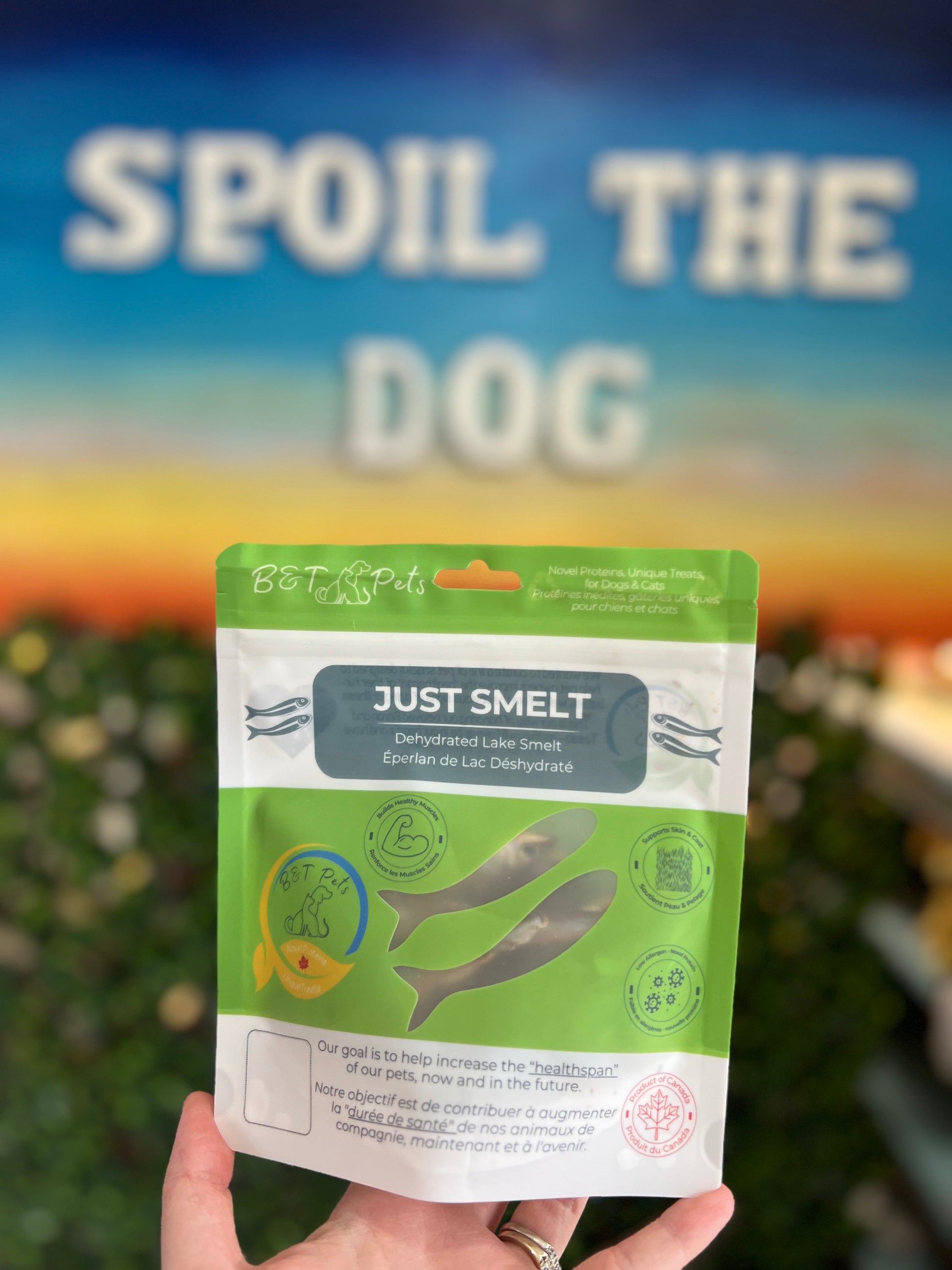 Hand holding a green package labeled 'Just Smelt' with a blurred background featuring 'Spoil the Dog'.