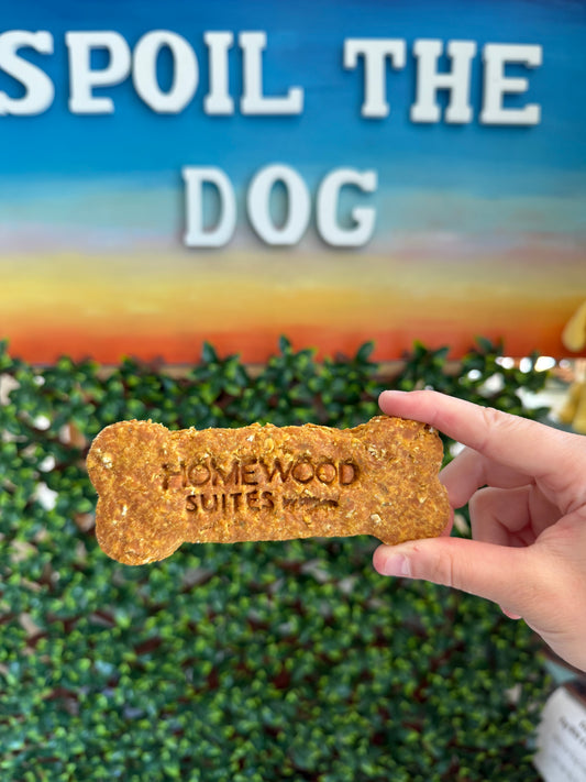 Dog treat shaped like a bone with 'Homewood Suites' branding, held by a hand against a blurred background.
