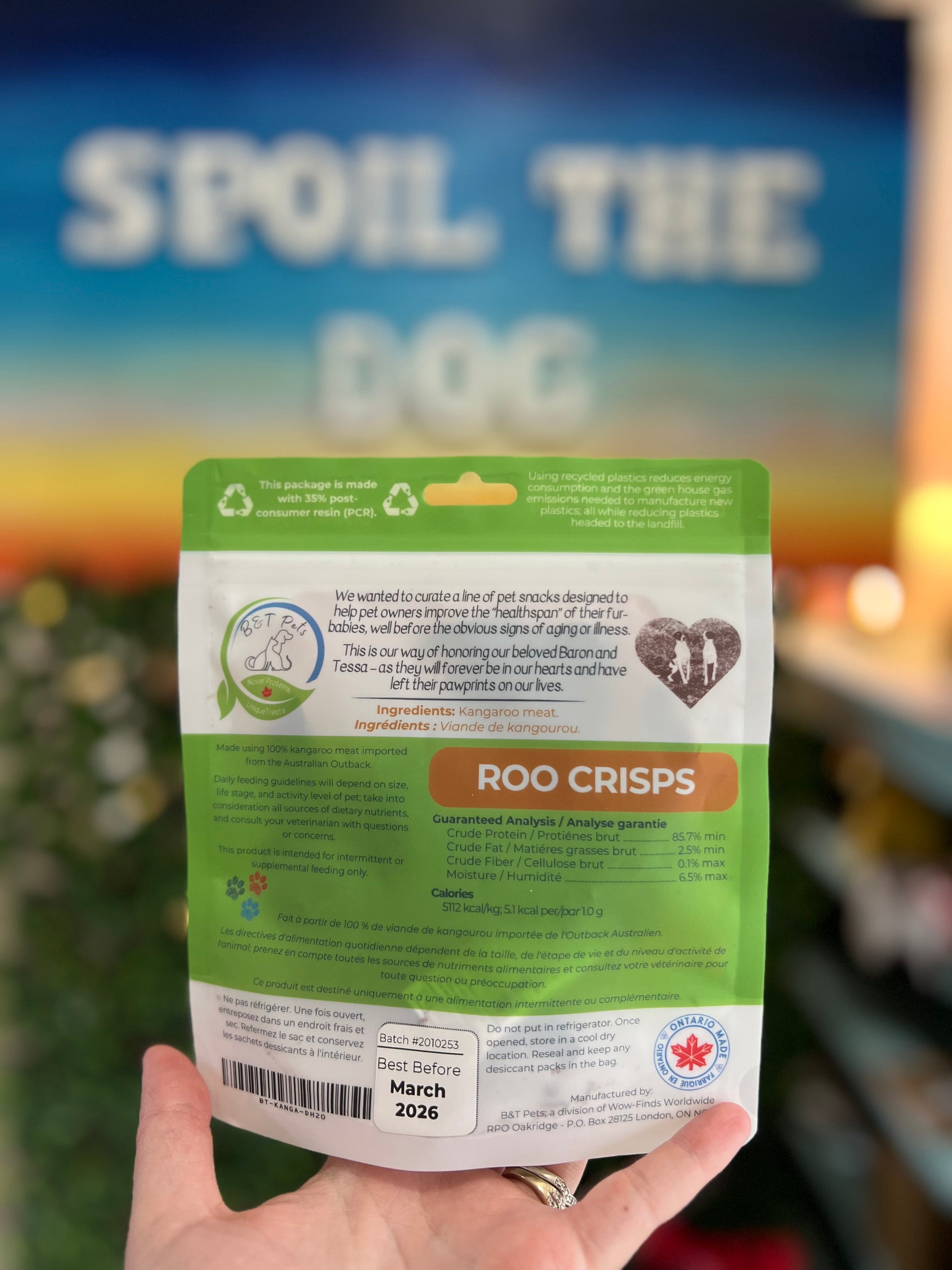 Hand holding a package of 'Roo Crisps' with a blurred background featuring 'Spoil the Dog'.
