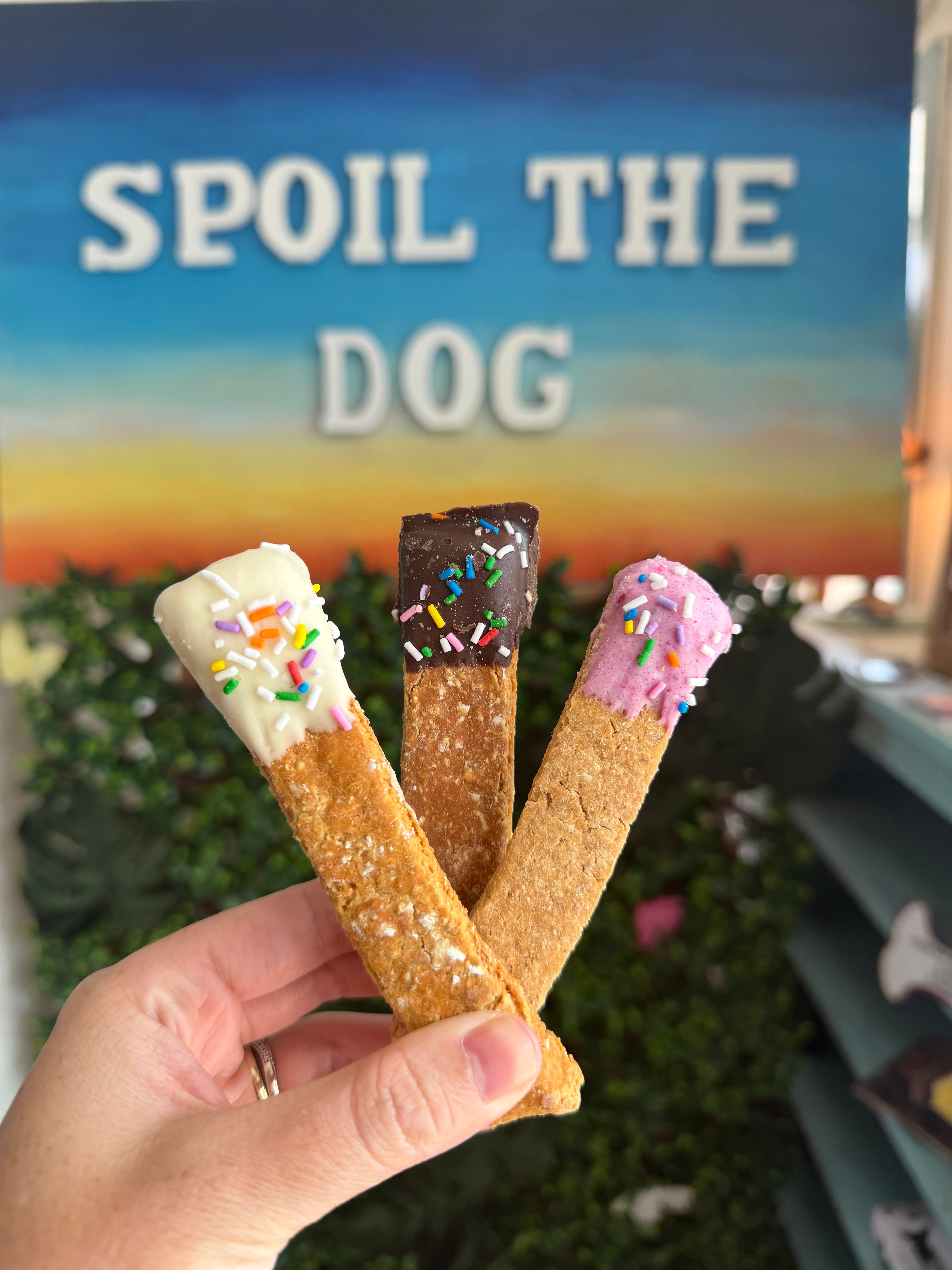 Three dog treats with colorful sprinkles and ice cream-style dips are held in a hand in front of a sign that reads 'SPOIL THE DOG'.