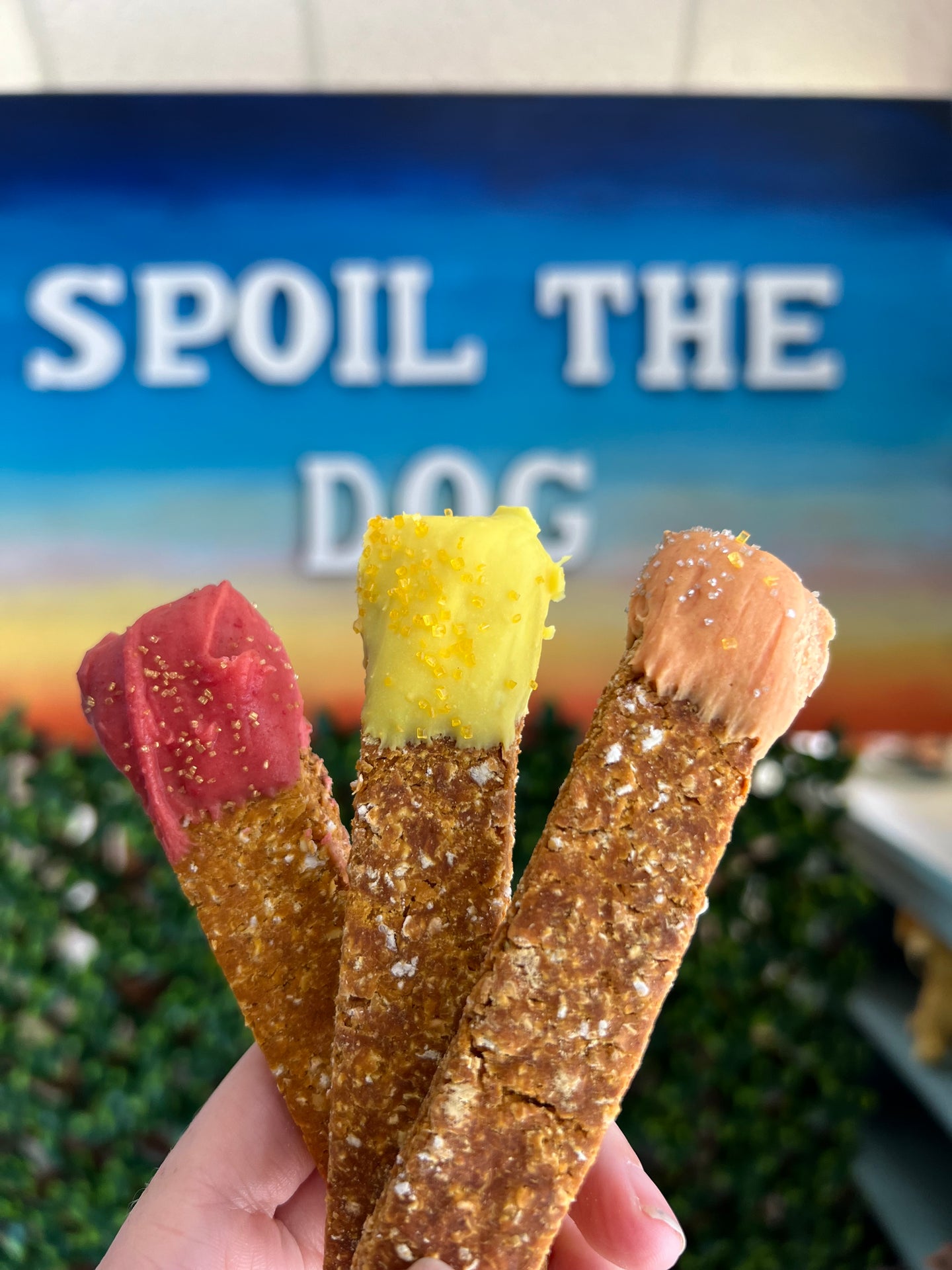 Two dog treats held by a hand with 'Spoil The Dog' sign in the background.