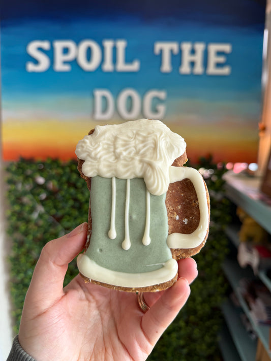 Hand holding a dog treat shaped like a green beer with 'spoil the dog' sign in the background