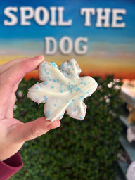 Hand holding a white snowflake dog treat with blue sprinkles in front of a 'Spoil the Dog' sign.