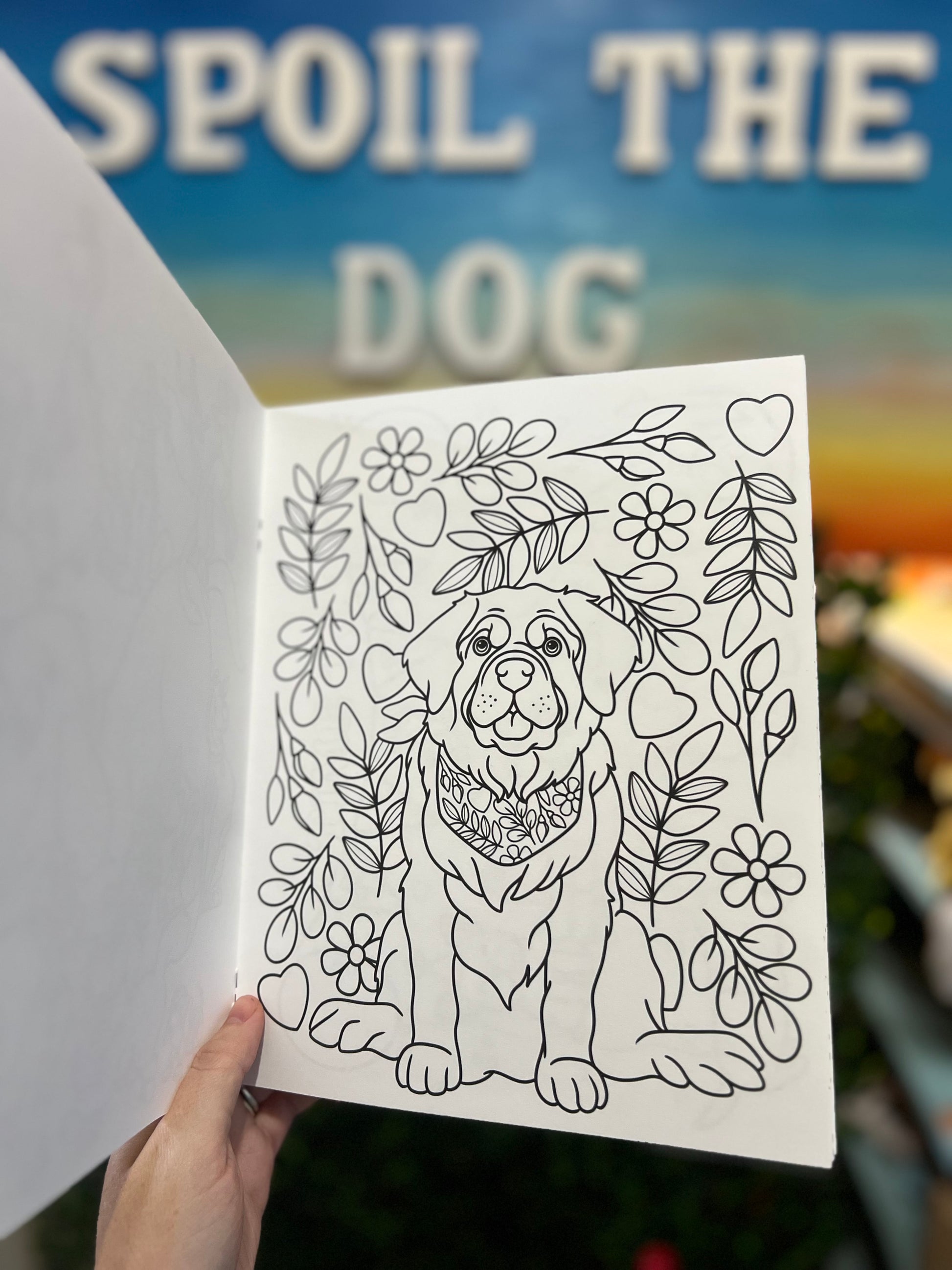 Coloring book page with a dog illustration surrounded by flowers and leaves, held against a blurred background with text.