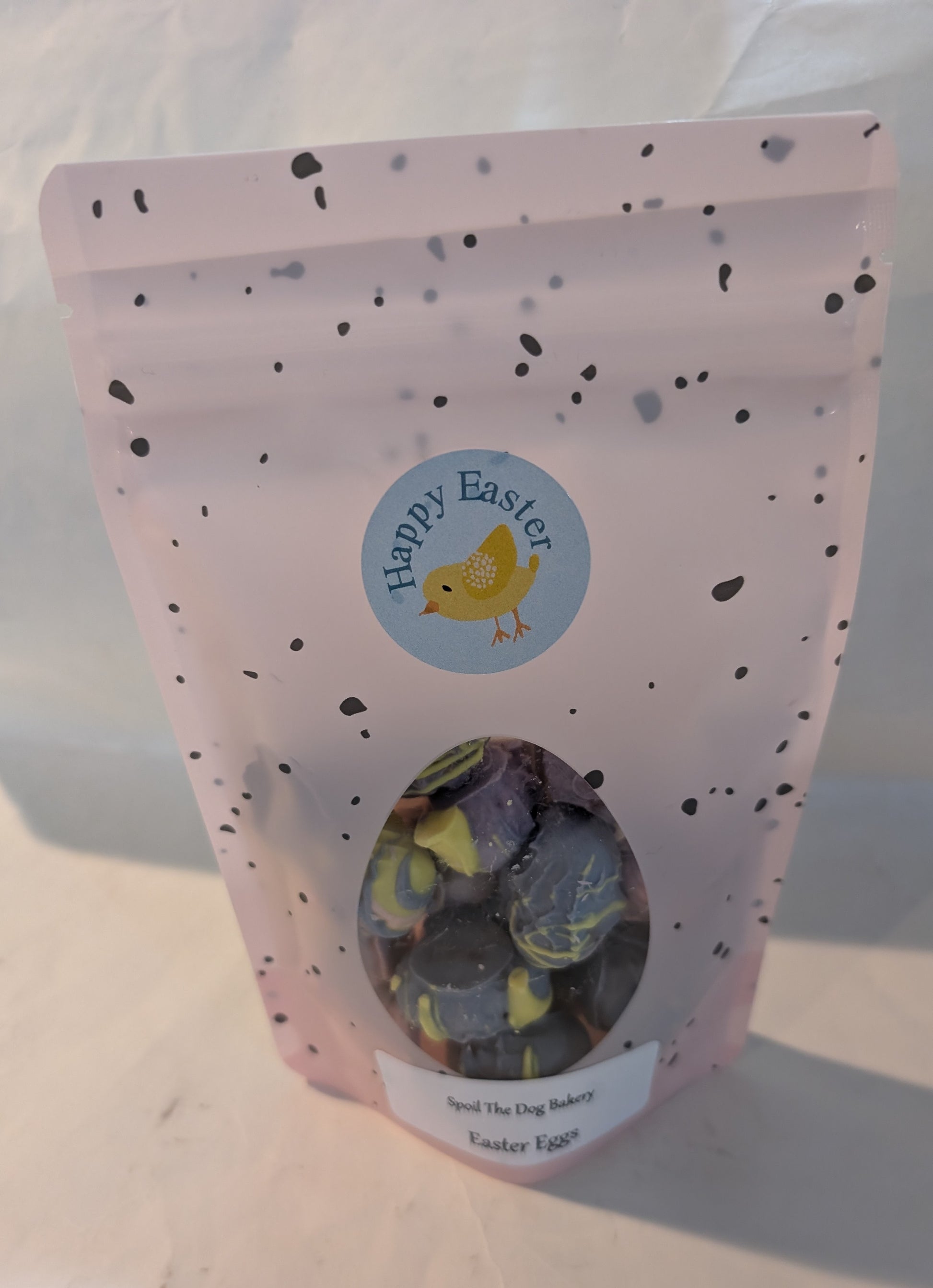 A sealed, pink treat bag labeled 'Happy Easter' with a transparent window revealing peanut butter eggs coated in carob and yogurt pieces for dogs.