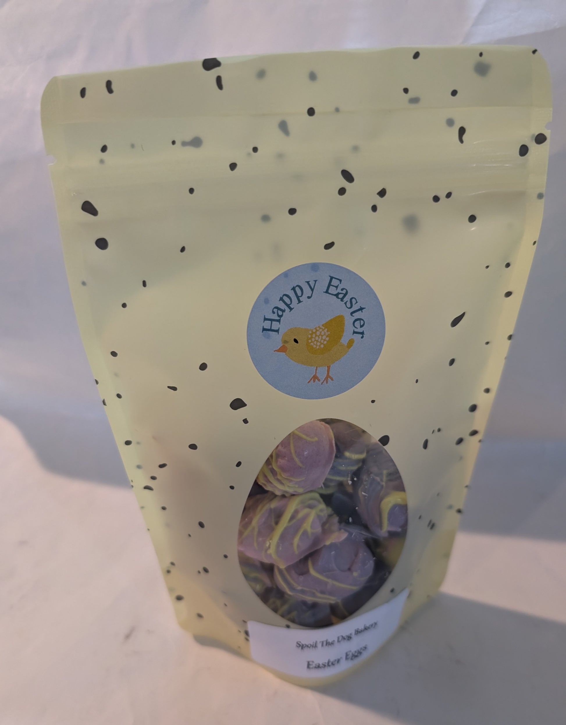 A sealed, yellow treat bag labeled 'Happy Easter' with a transparent window revealing peanut butter eggs coated in carob and yogurt pieces for dogs.
