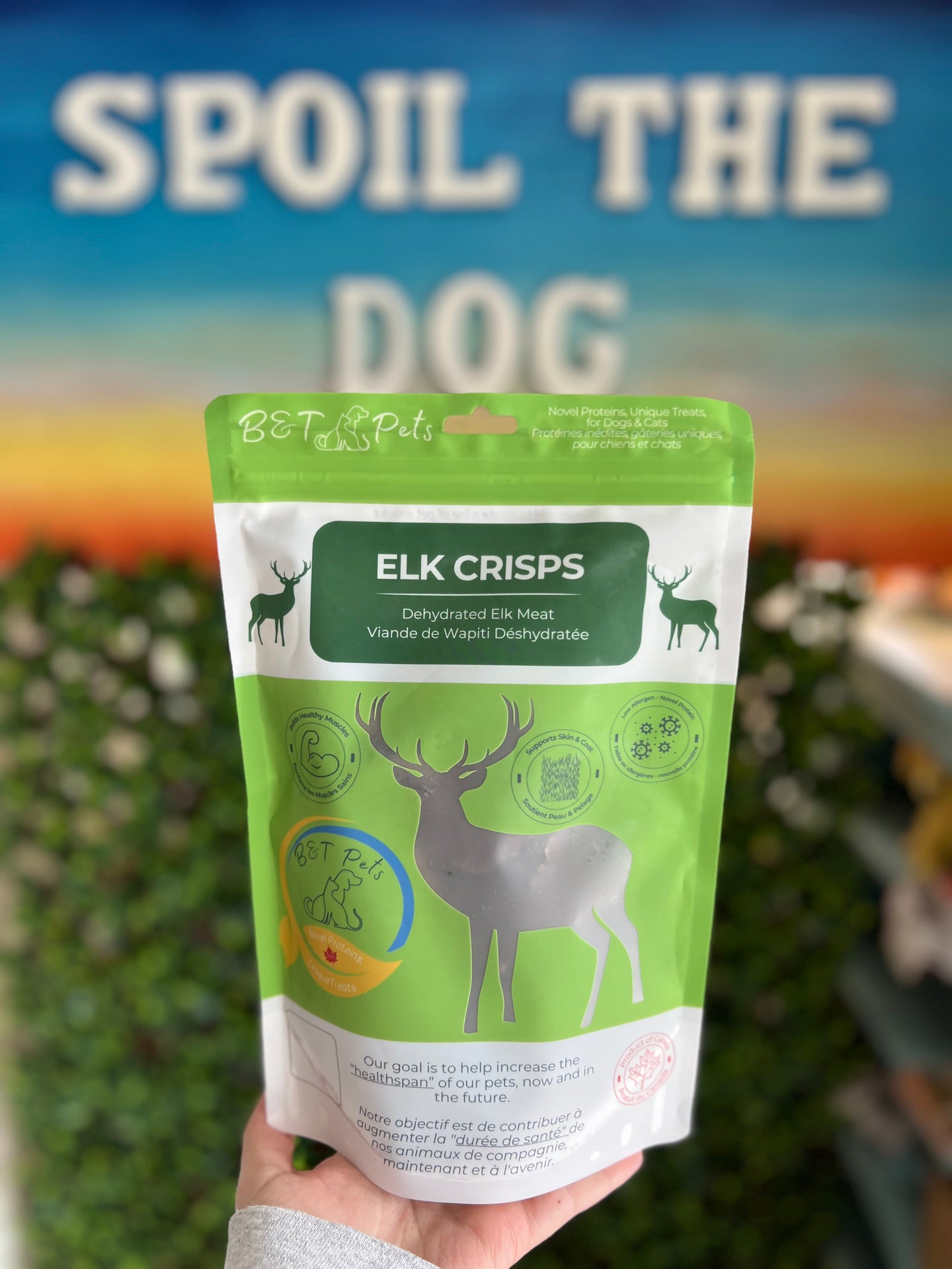 Hand holding a package of Elk Crisps with a blurred background featuring 'SPOIL THE DOG' text.