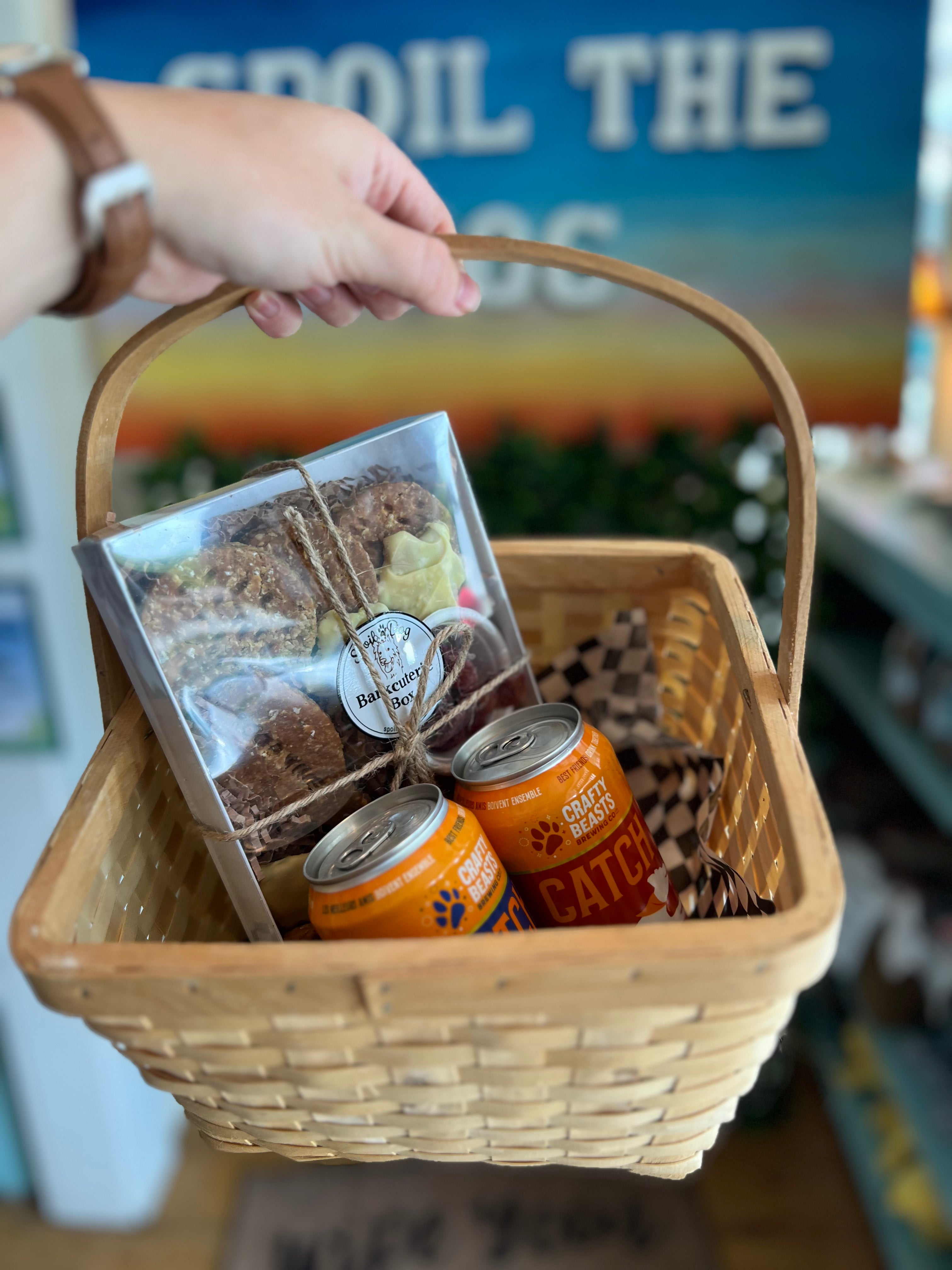 Wicker basket with pet supplies including dog beer cans and a barkcuterie box, held by a person.