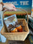Wicker basket with pet supplies including dog beer cans and a barkcuterie box, held by a person.