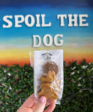 Packaged dog treats held in front of a 'Spoil the Dog' sign with a scenic background.