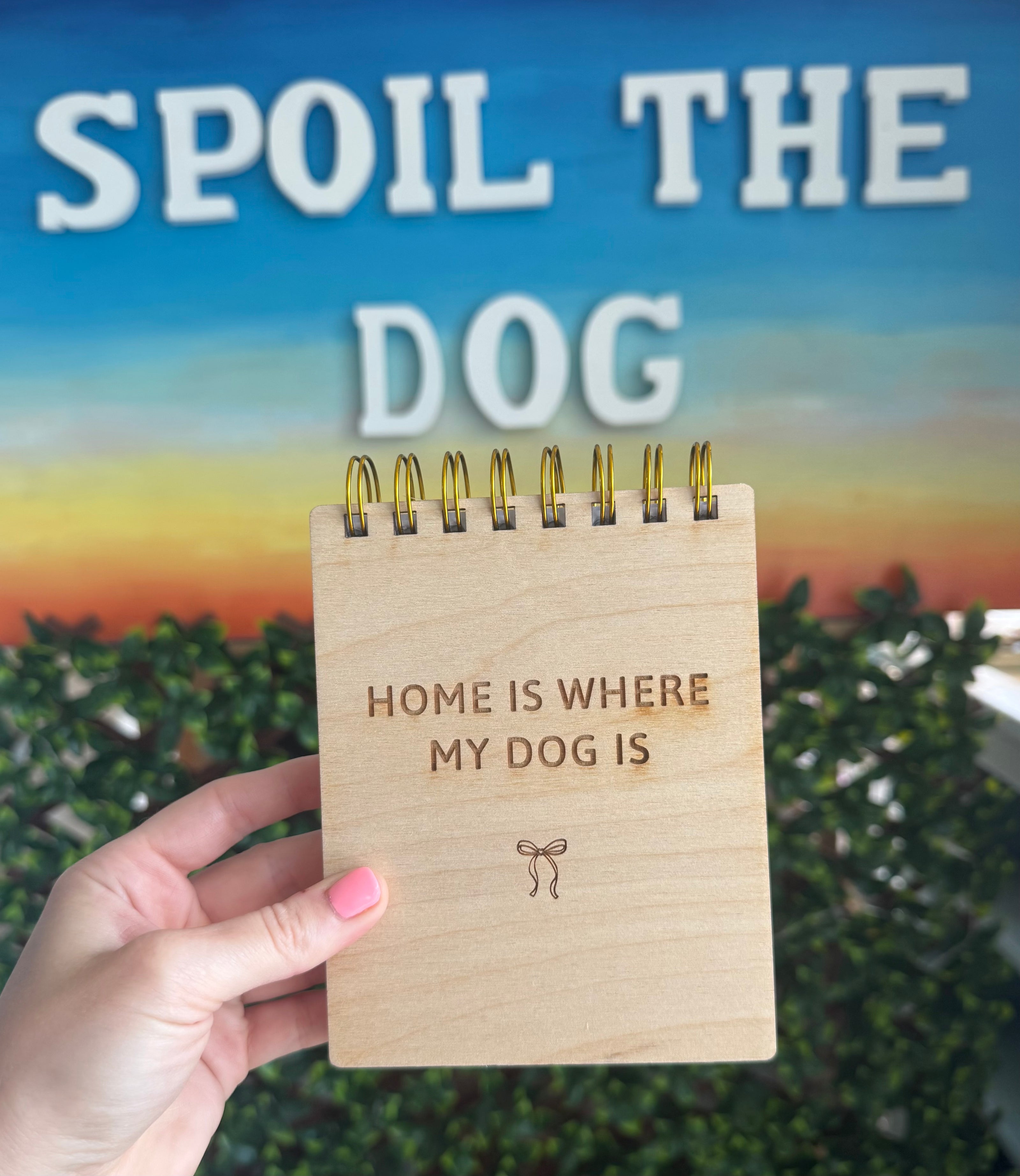 A hand holds a small wooden spiral-bound notepad that reads 'HOME IS WHERE MY DOG IS' with a small bow engraving. Behind it is a large printed sign reading 'SPOIL THE DOG' over a blue sky and green hedge background.