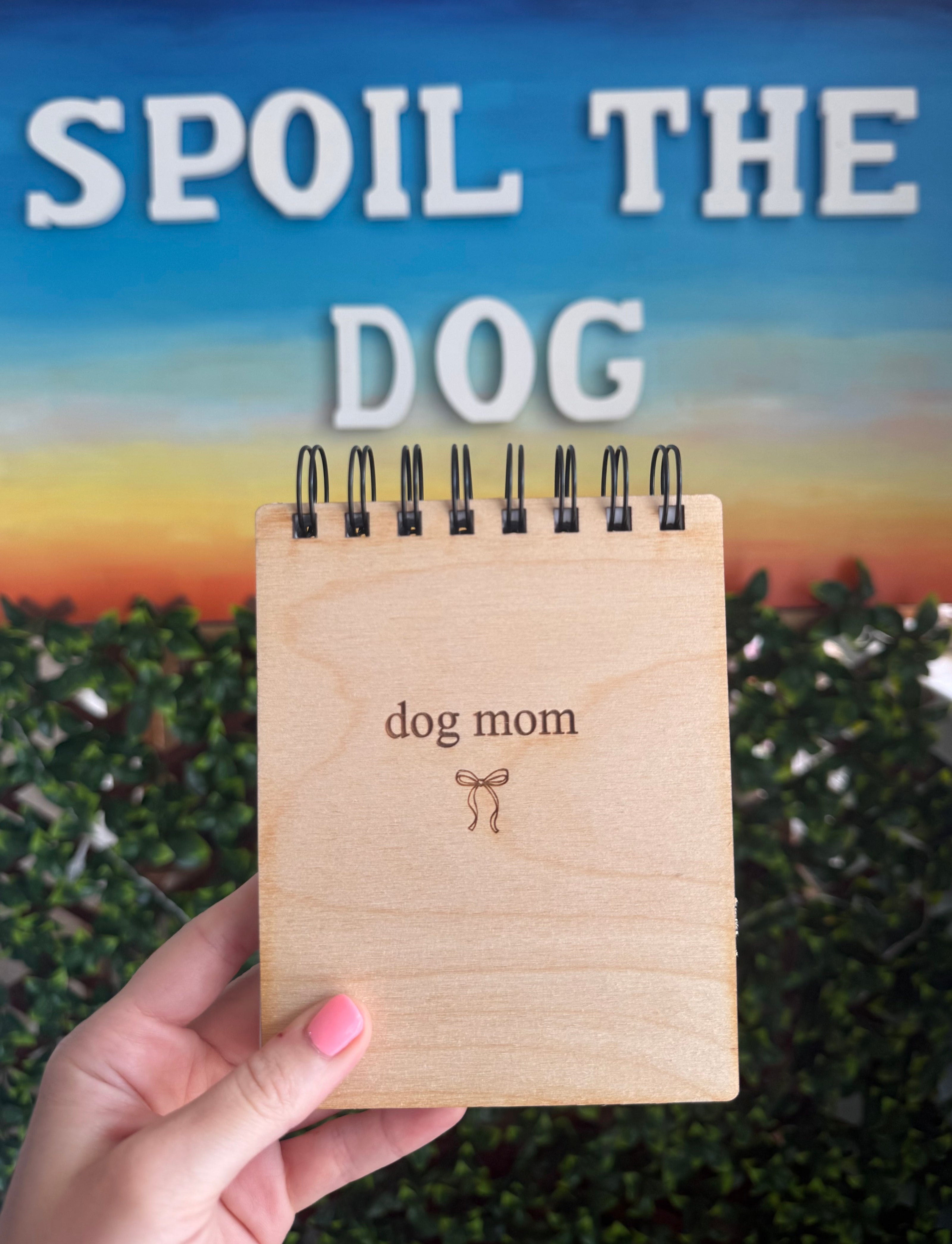A hand holds a small wooden spiral-bound notepad that reads 'DOG MOM' with a small bow engraving. Behind it is a large printed sign reading 'SPOIL THE DOG' over a blue sky and green hedge background.