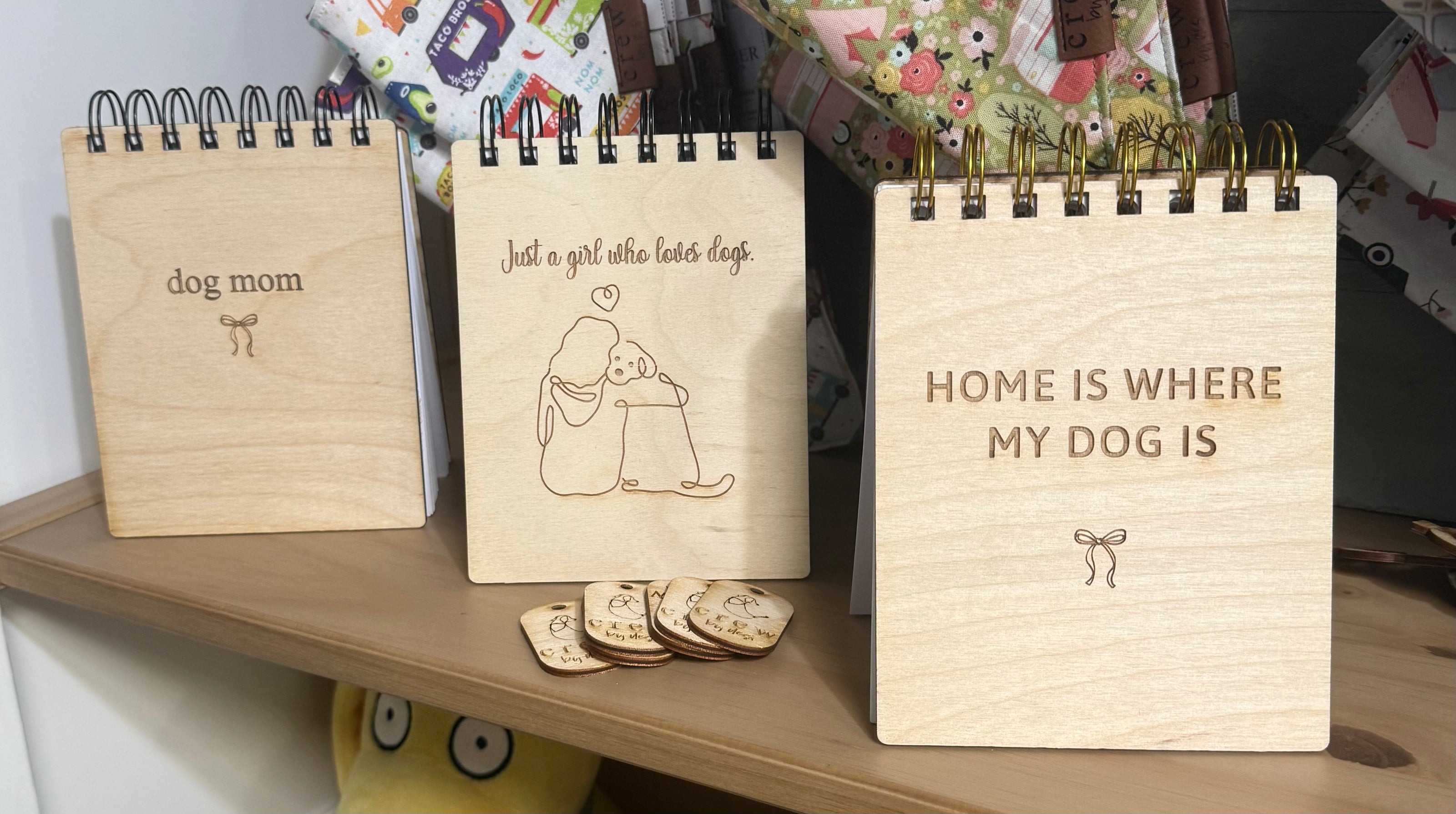 Three small wooden spiral-bound desk pads or mini notebooks displayed on a shelf, each engraved with a dog-themed phrase: 'dog mom,' 'Just a girl who loves dogs,' and 'HOME IS WHERE MY DOG IS.'.