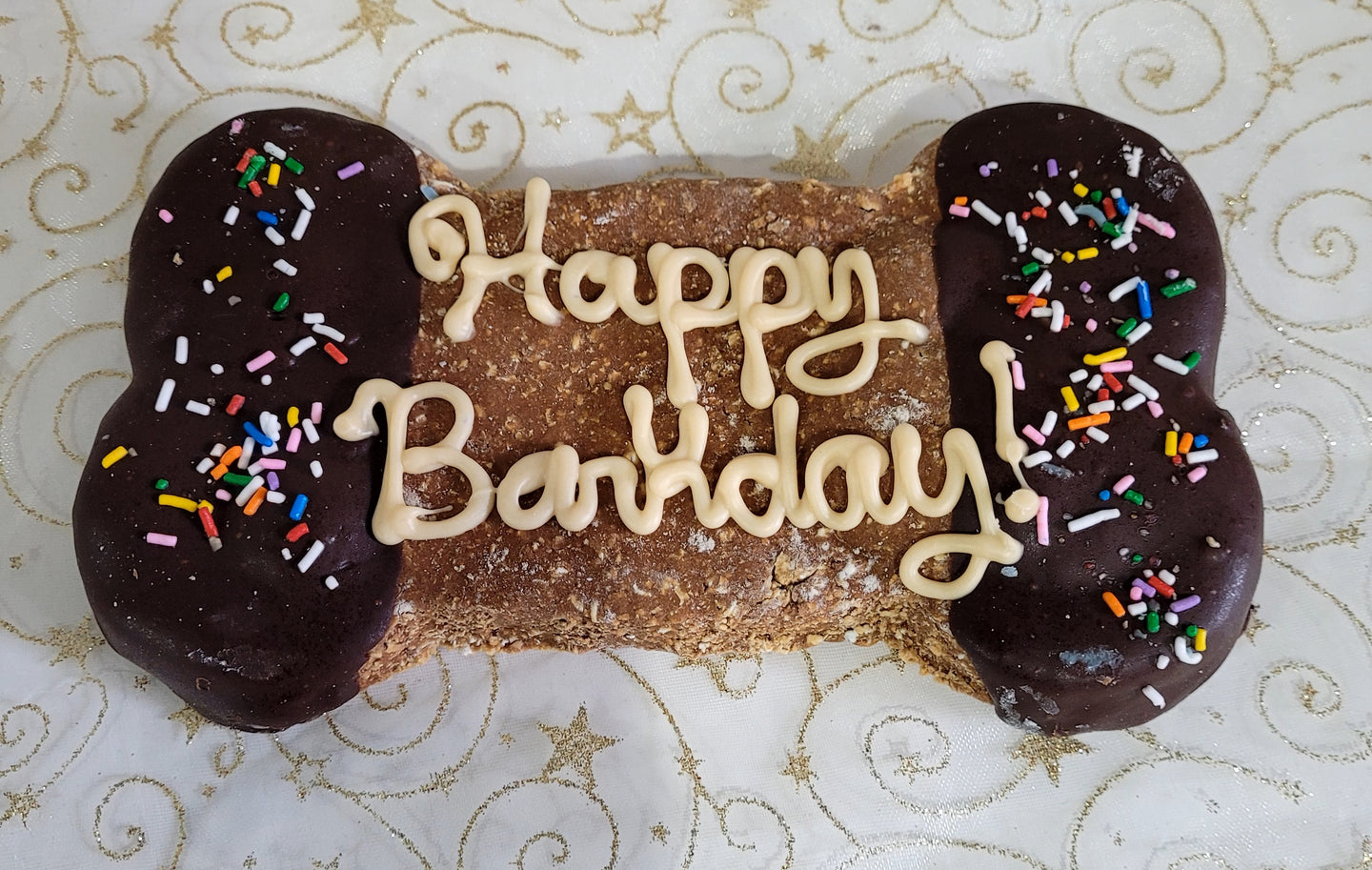 A jumbo bone-shaped dog treat with 'Happy Barkday' text and sprinkles on the bone's top edge, placed on a wooden surface.