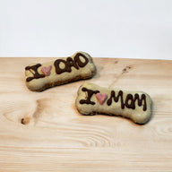 Two bone-shaped dog treats with 'I LOVE' and 'Dad' or 'Mom' written on them, placed on a wooden surface.