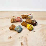 A selection of dog treats in the shape of mini bones, with varying colors and sprinkles, displayed on a wooden surface.