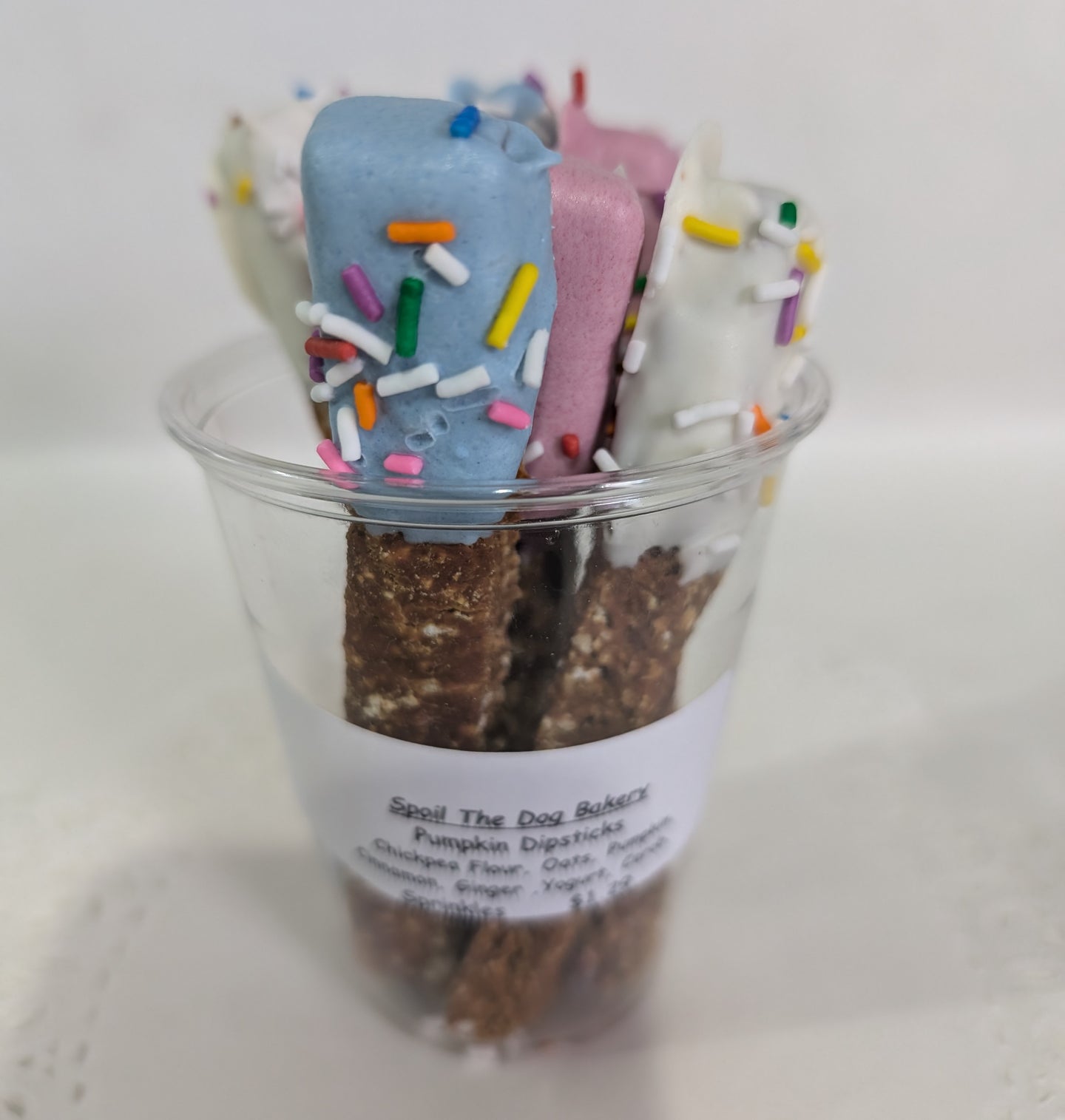 A clear plastic cup containing dog treats. The treats are dipped in various coatings, such as blue and white icing, and are sprinkled with colorful toppings.