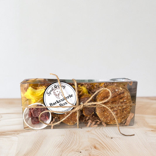 A dog treat barkcuterie box containing various natural dog treats such as 'crackers', 'cheese' diamonds, freeze dried strawberries and a squirrel shaped treat, displayed on a wooden surface.