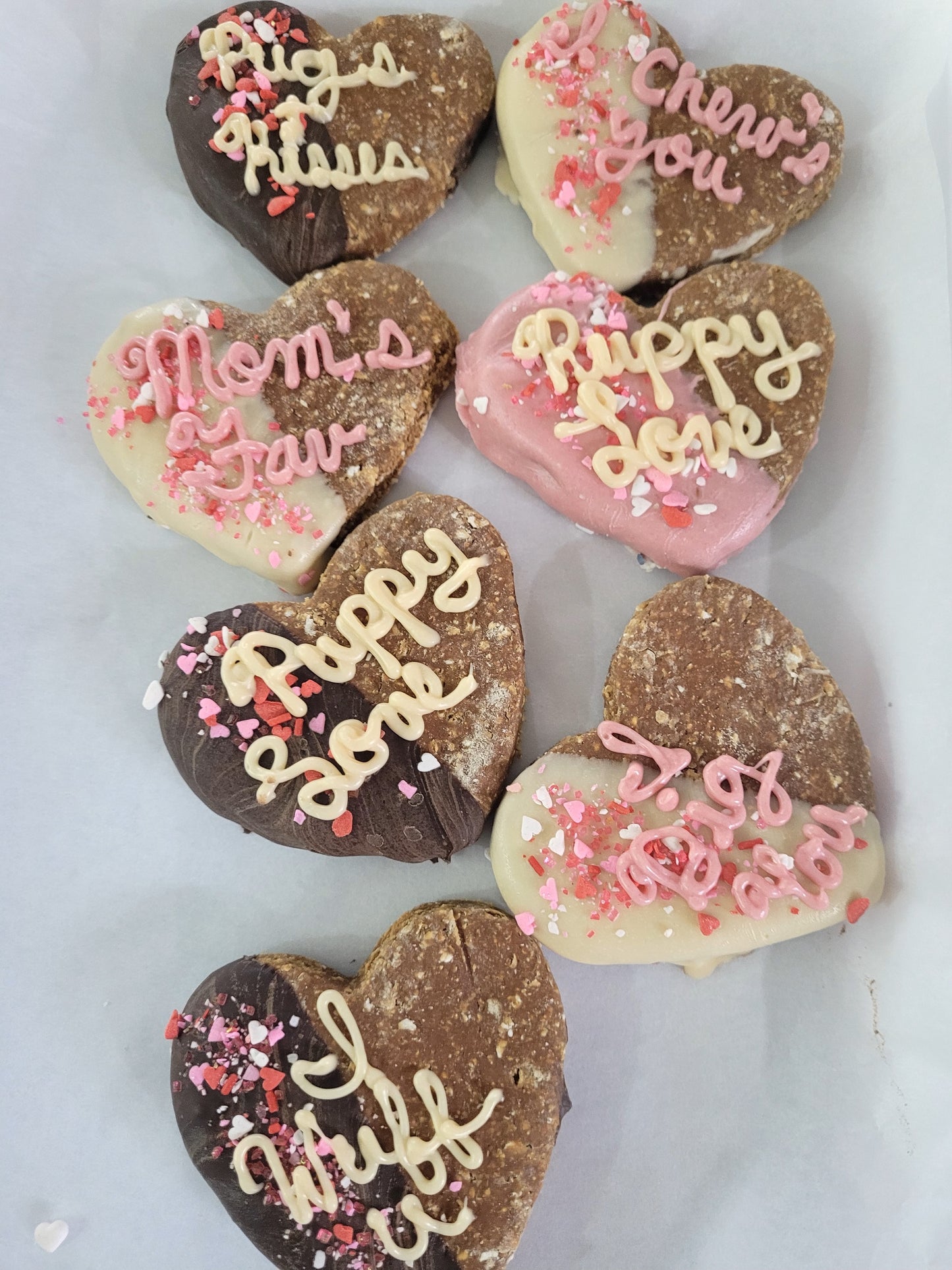 A variety of heart-shaped dog treats with different messages such as 'Happy Valentine's Day' and 'I love you' written on them, dipped in yogurt and carob.