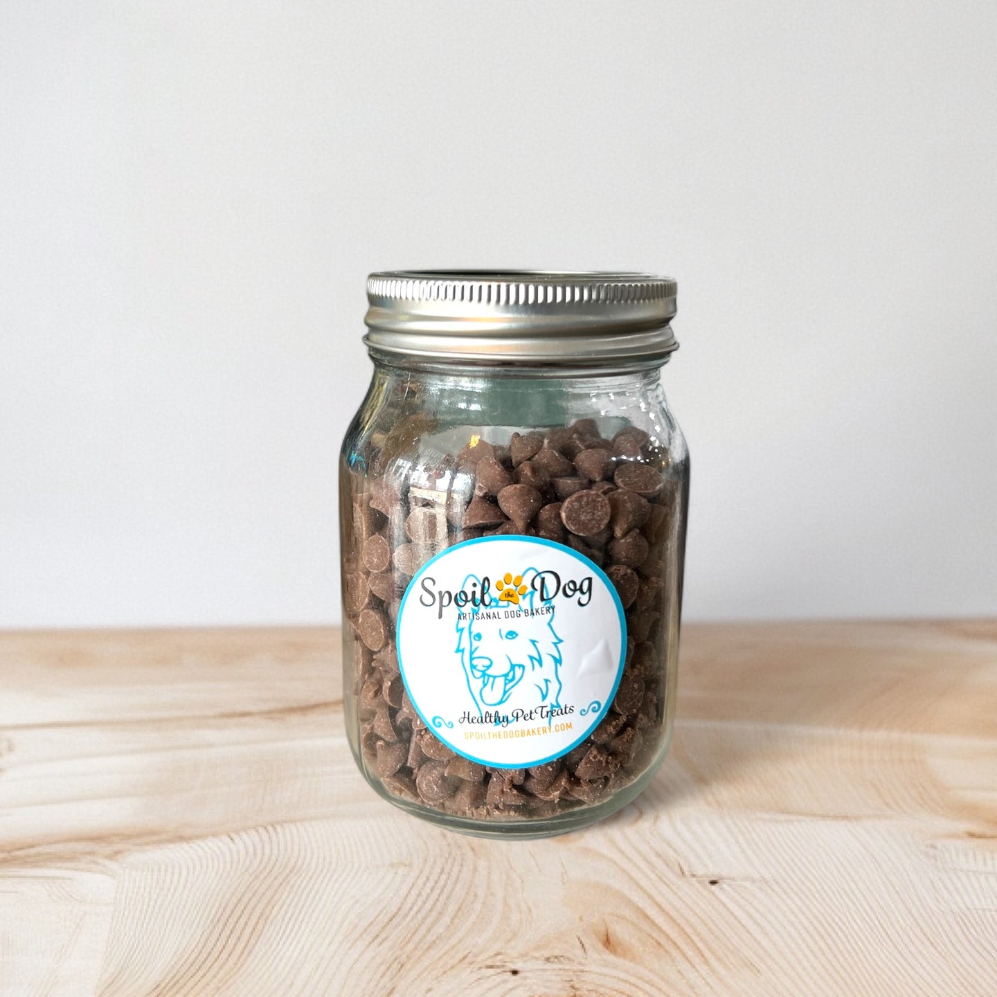 A glass jar filled with carob chips dog treats, labeled with 'Spoil the Dog Bakery' on a wooden surface.