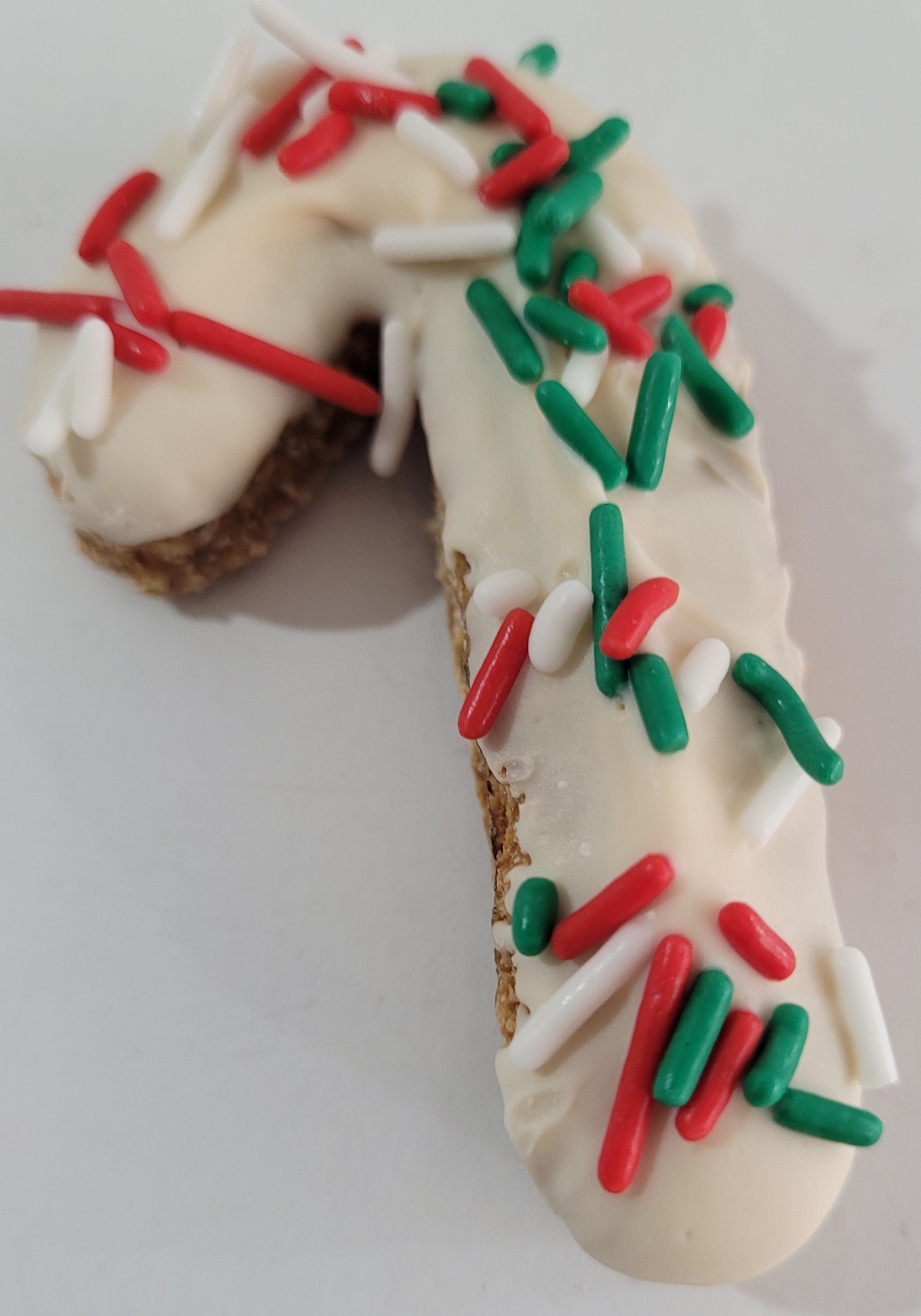 A peanut butter candy cane-shaped dog treat with chickpea flour, oats, peanut butter, yogurt, and sprinkles, decorated with red and green sprinkles on a white background.