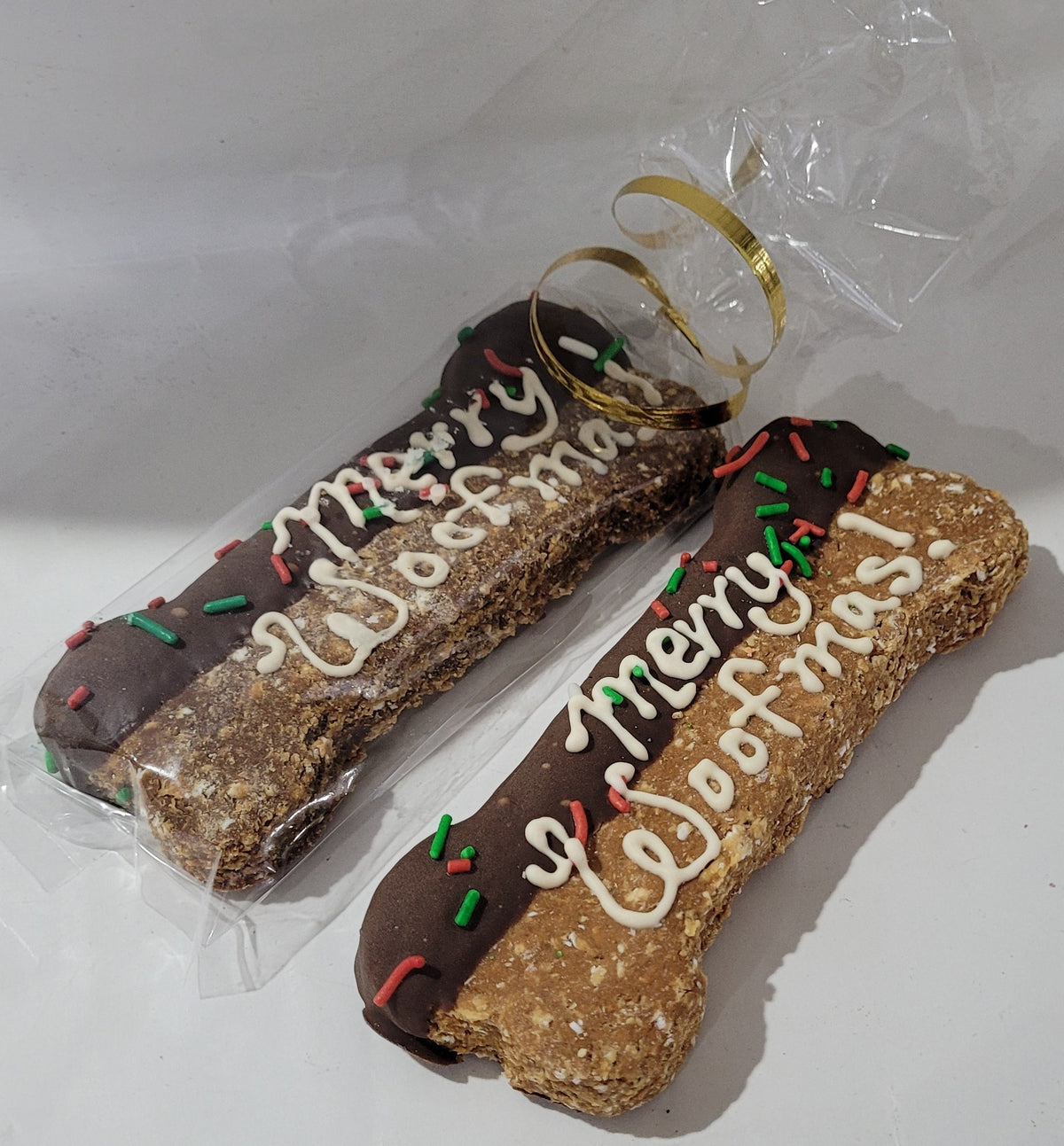 Two decorated dog treats with 'Merry Woofmas' and 'Woofmas' text, one with a golden yellow bow on top, the other with red and green accents. Both are packaged in clear plastic with a white background.