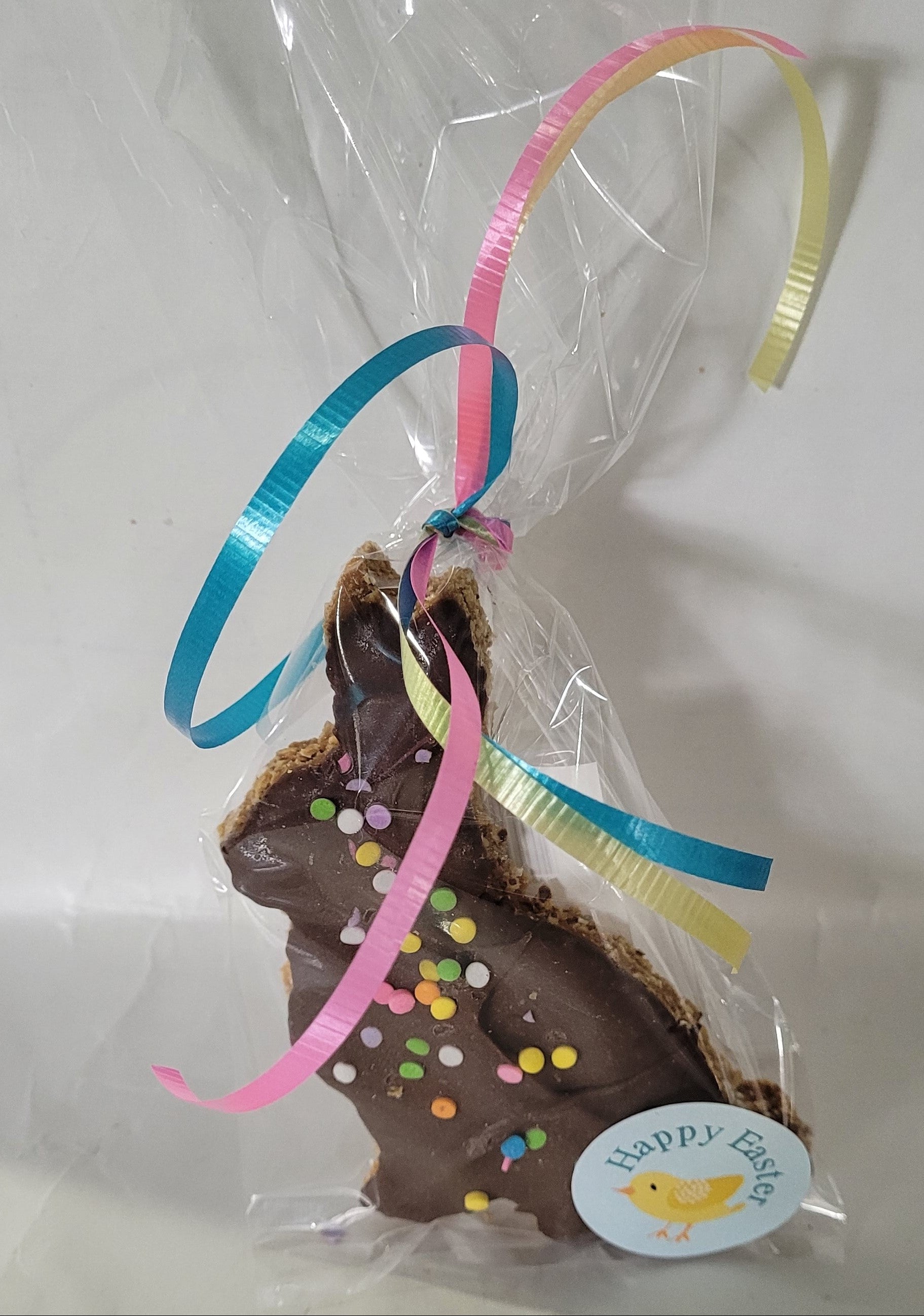 A carob-coated dog treat shaped like a rabbit, decorated with a pink and yellow ribbon and multicolored sprinkles, presented in a clear packaging with a 'Happy Easter' label.